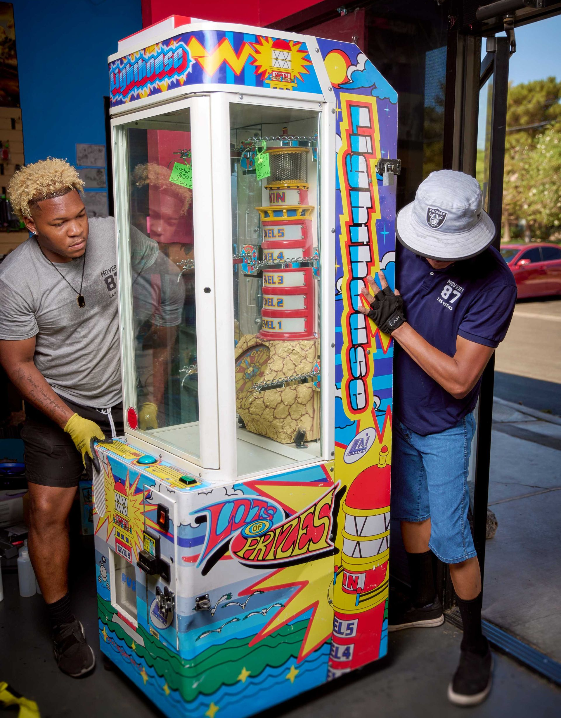 Two people moving a claw machine through a doorway. The machine is colorful with toys inside.