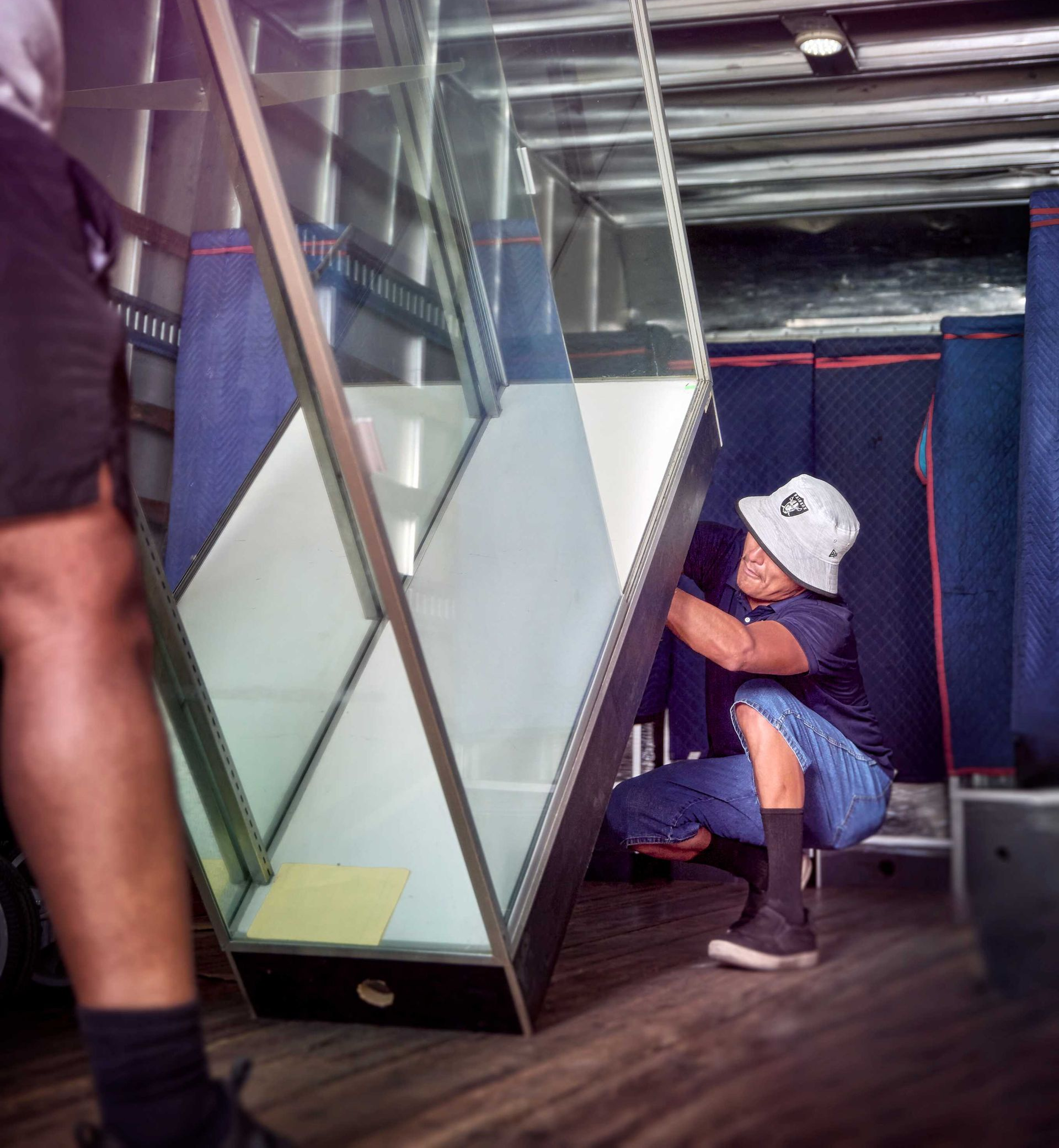 Two people lift a large, rectangular glass shelf unit inside a vehicle. One wears a hat.