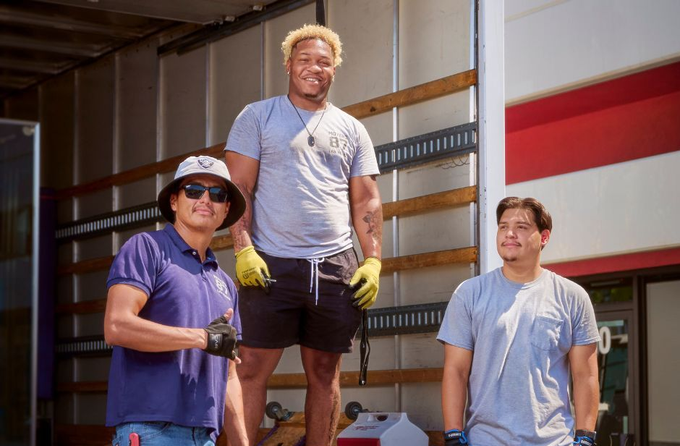 Three movers standing in front of a truck, smiling. One is wearing a hat, another is on a lift, and all are in work clothes.
