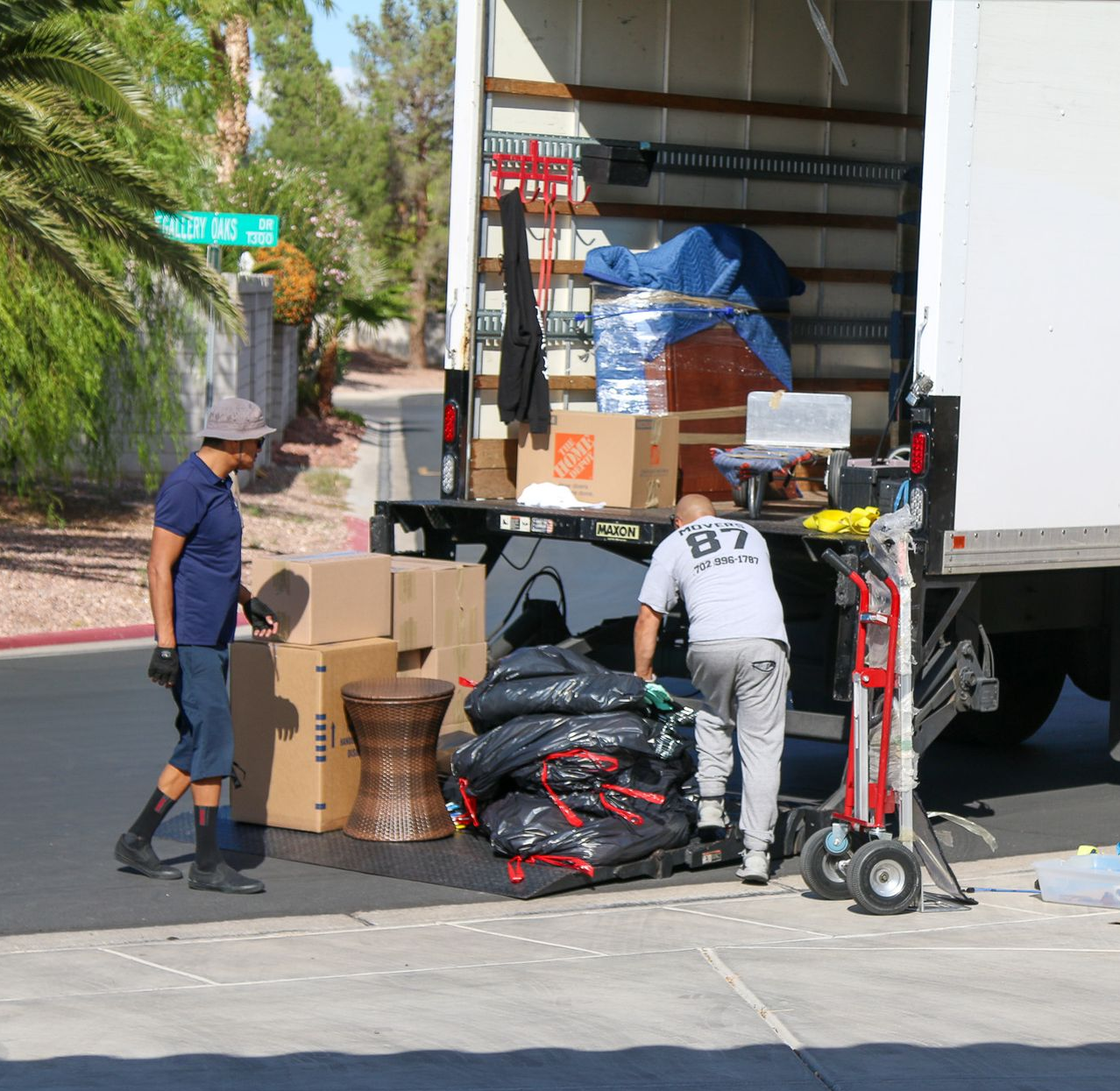Two people loading a moving truck on a sunny street, boxes and furniture on the ground, palm tree in background.