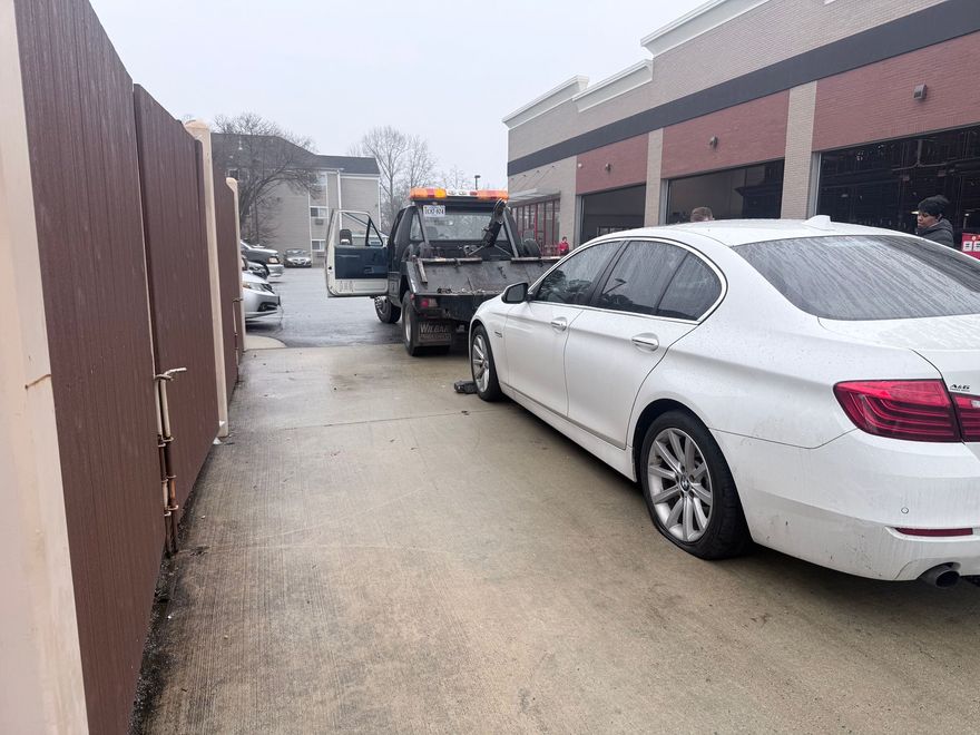 A white sedan parked on a concrete lot next to a brown wooden fence, with a tow truck positioned in front of it.