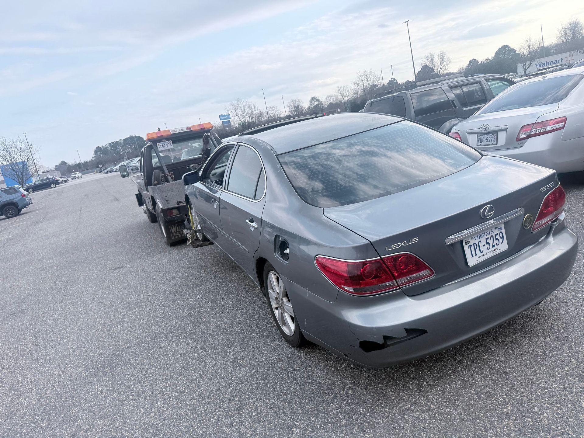 A grey Lexus sedan is being towed by a tow truck in an outdoor parking lot under a cloudy sky.