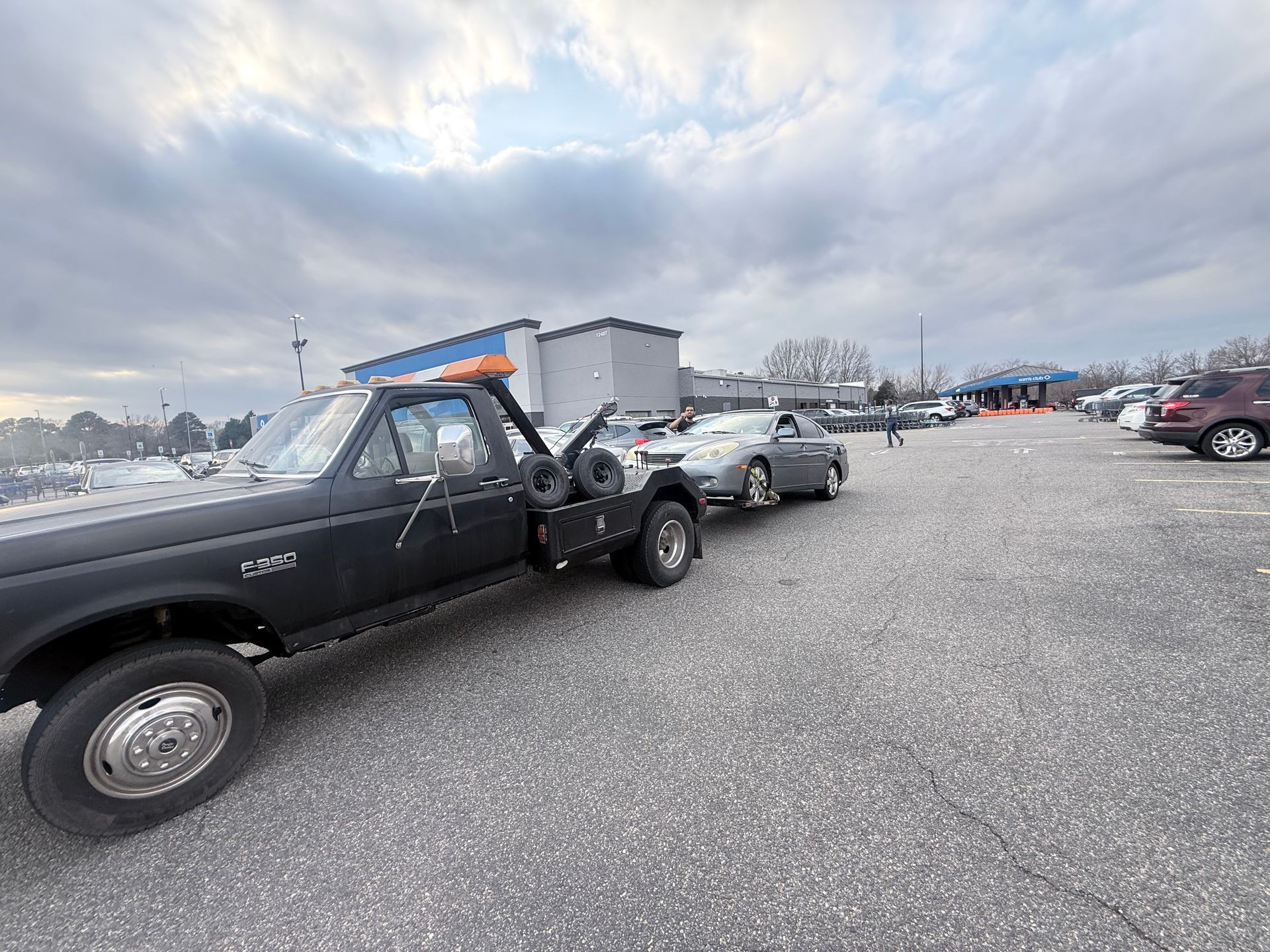 A black tow truck with an orange light bar on top is towing a silver sedan across a large, paved parking lot.