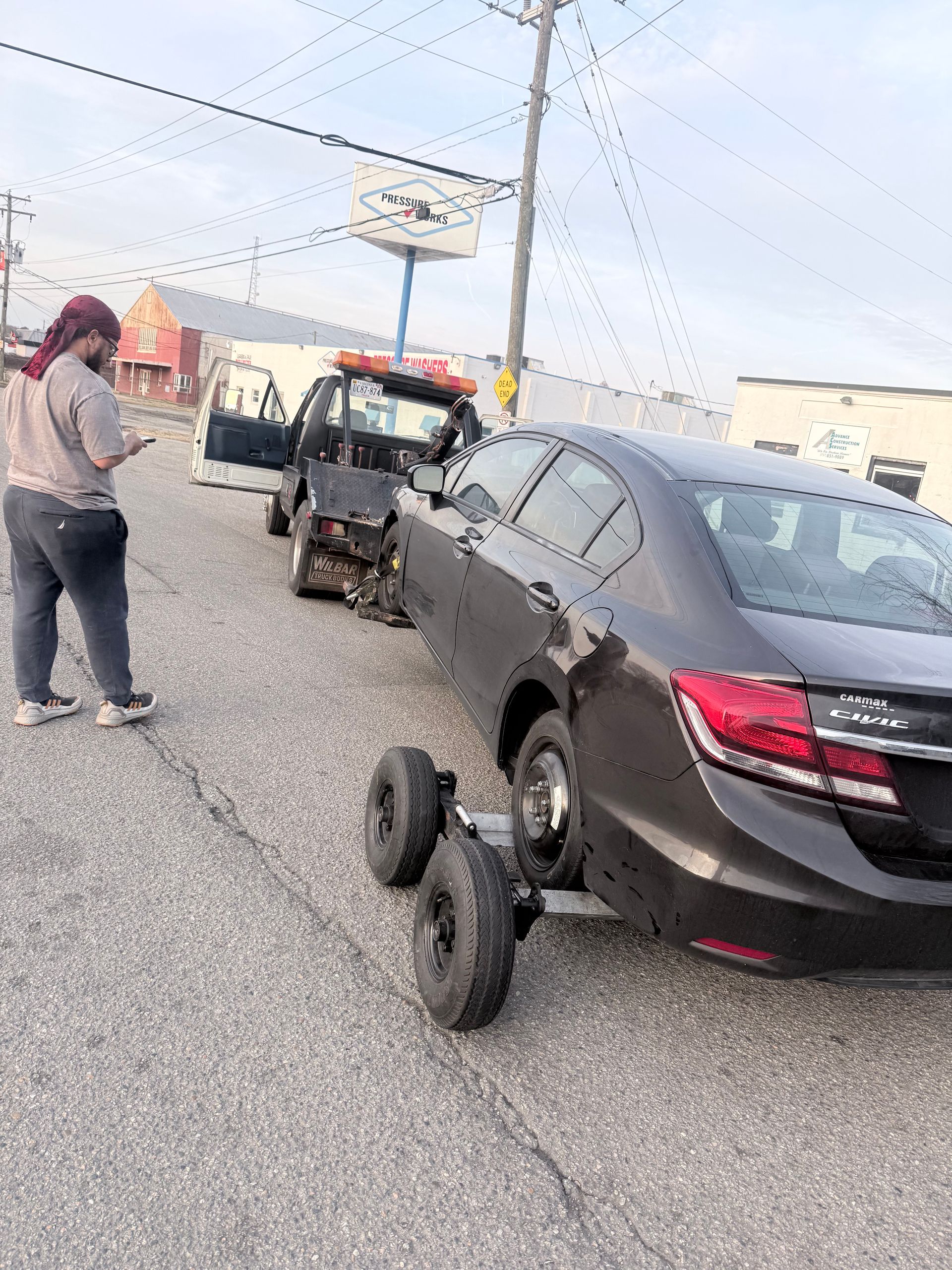 A person stands near a dark sedan being towed with dollies on its rear tires on an asphalt road.