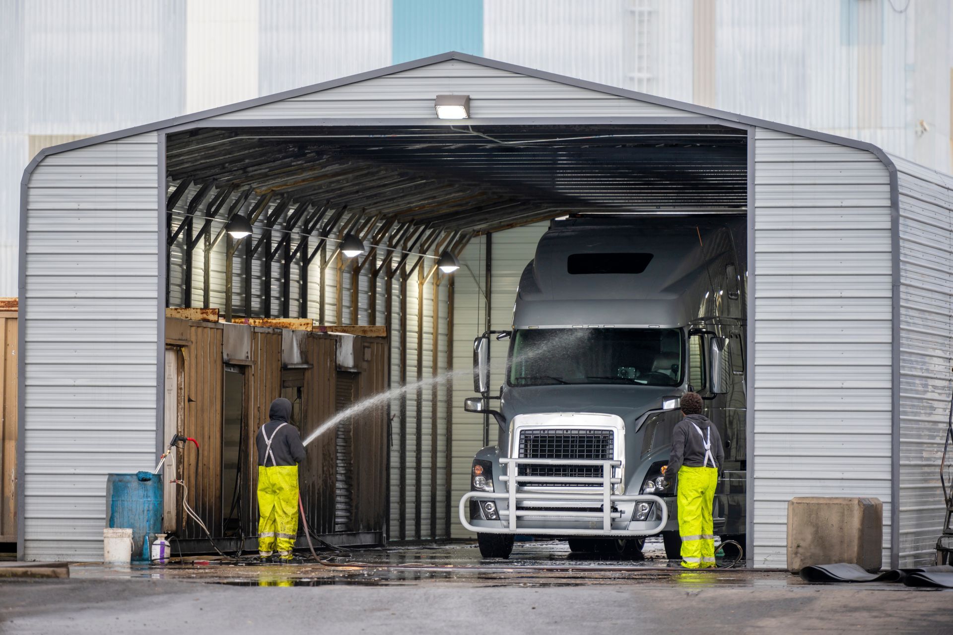 Two workers in yellow reflective pants wash a gray semi-truck inside an open metal-sided bay.