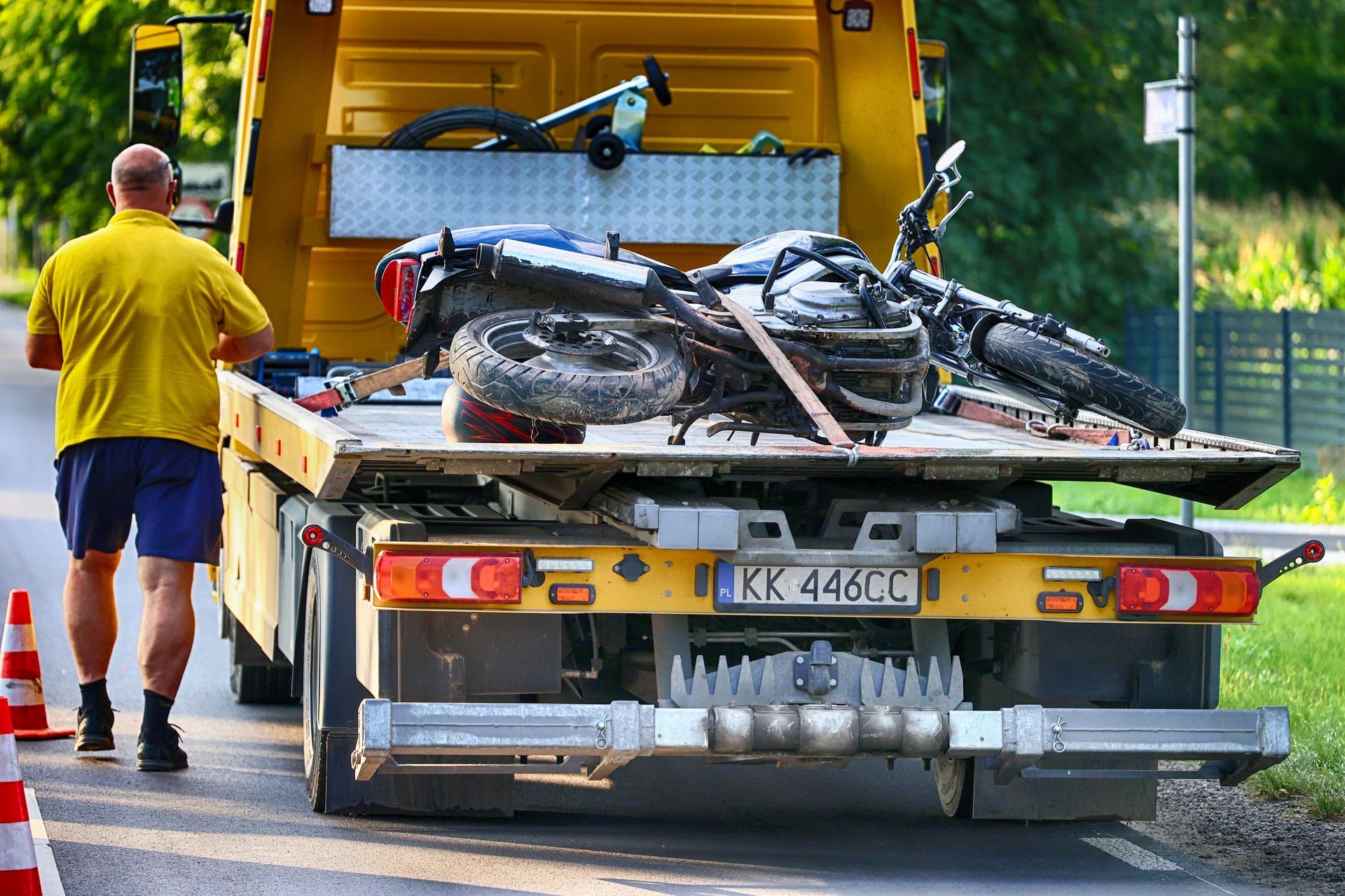 A flatbed tow truck carries a motorcycle on a road, with a person standing nearby in a yellow shirt and blue shorts.