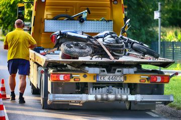 A flatbed tow truck carries a motorcycle on a road, with a person standing nearby in a yellow shirt and blue shorts.