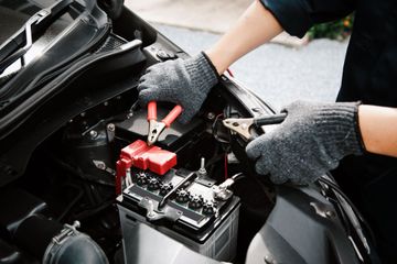 A person wearing work gloves connects jumper cables to a car battery under the hood of a vehicle.