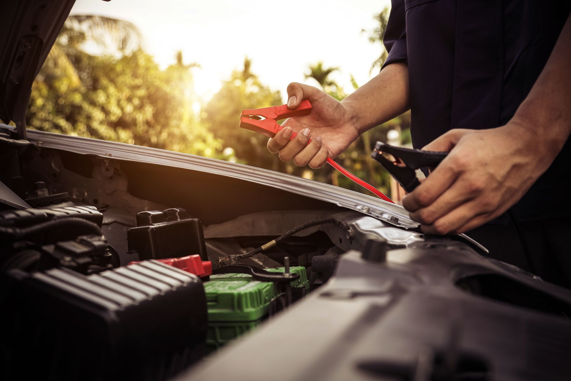 Hands holding red and black jumper cables over a car engine with a green battery in a sunny, outdoor setting.