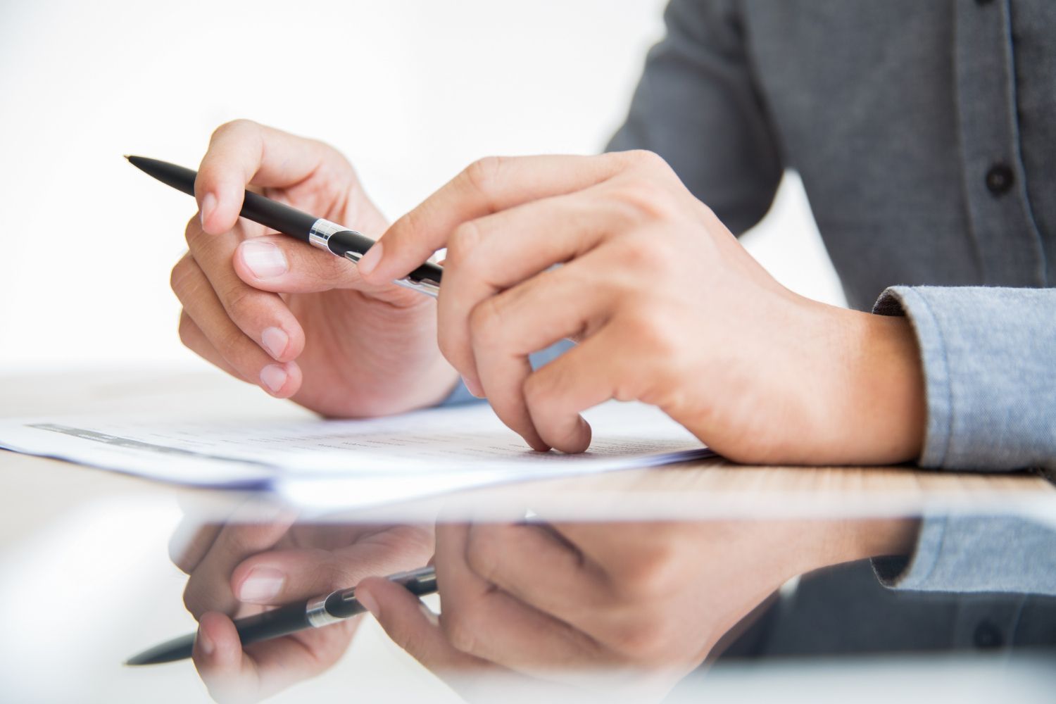 Person in gray shirt holding pen, fingers tapping on a document at a white table.