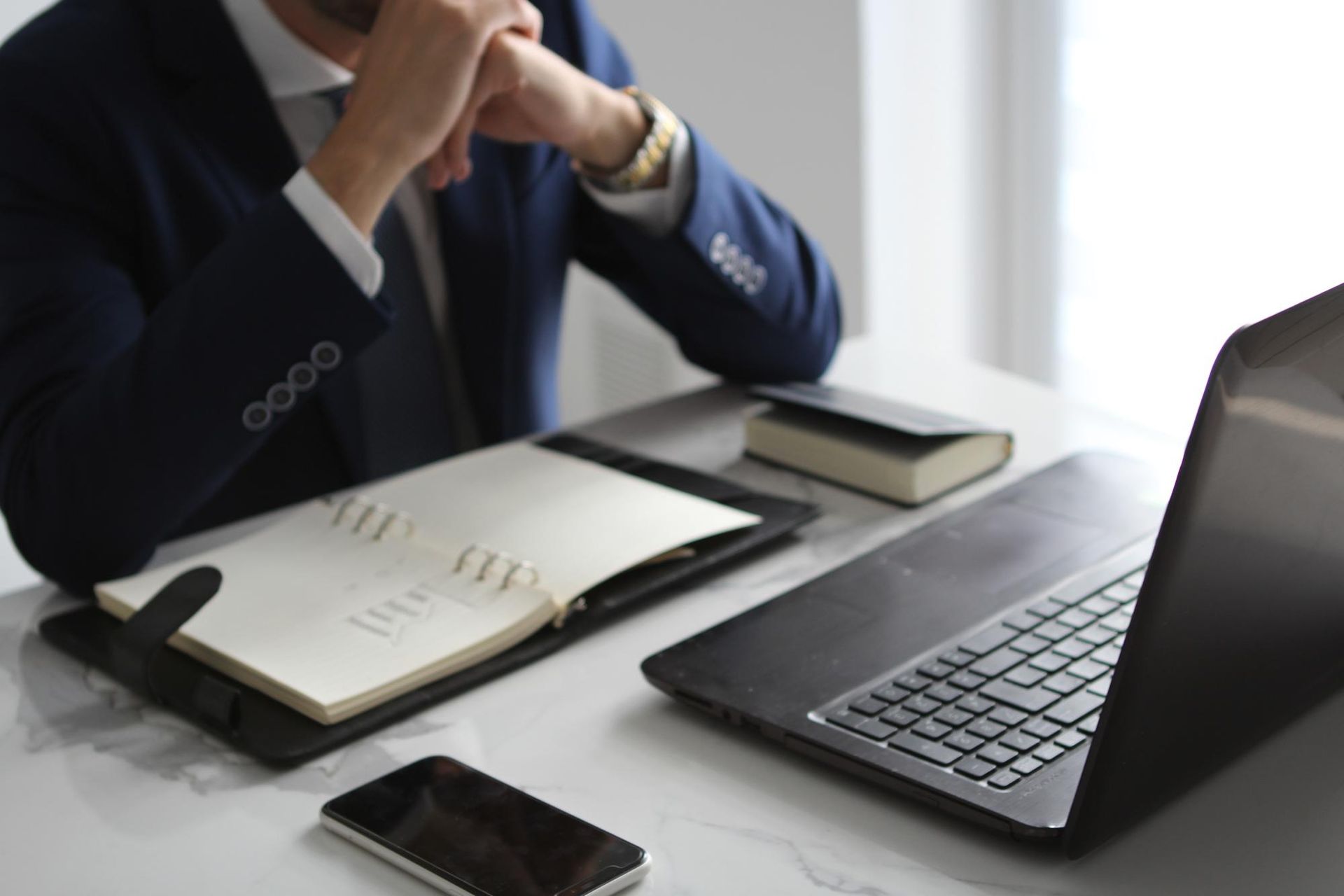 Man in suit with head in hands, at desk with laptop, notebook, and phone.