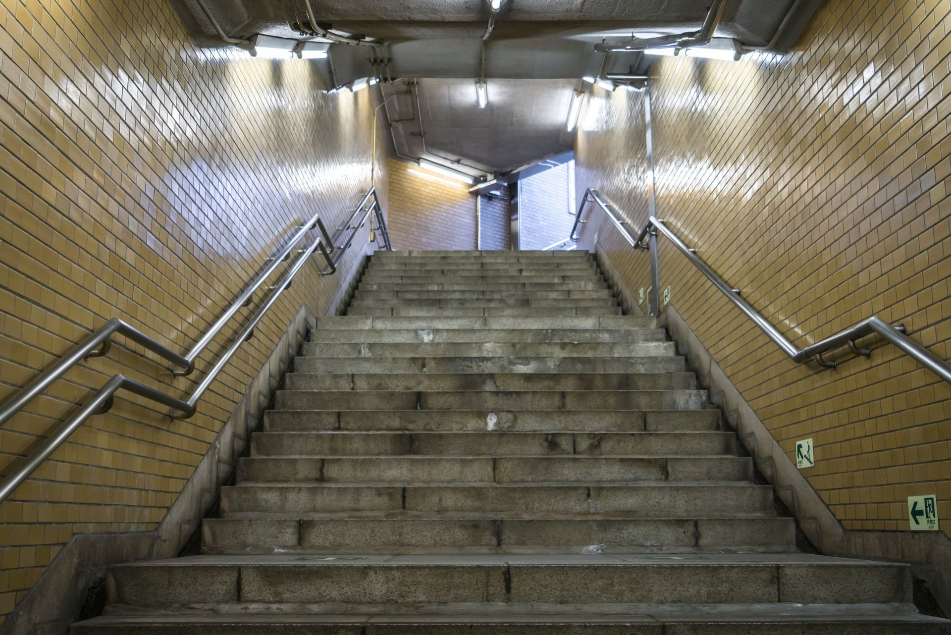 Staircase leading upwards in a subway tunnel; yellow tiled walls, metal handrails, and dim lighting.