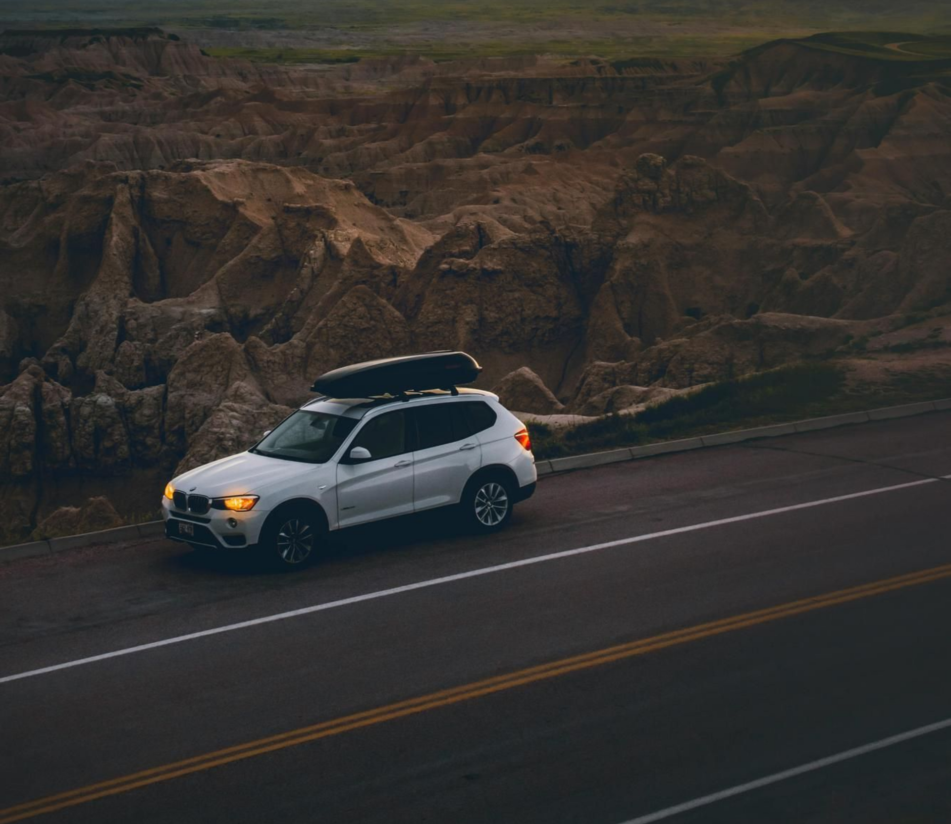 White SUV with a cargo box drives on a road next to rugged, brown rock formations.