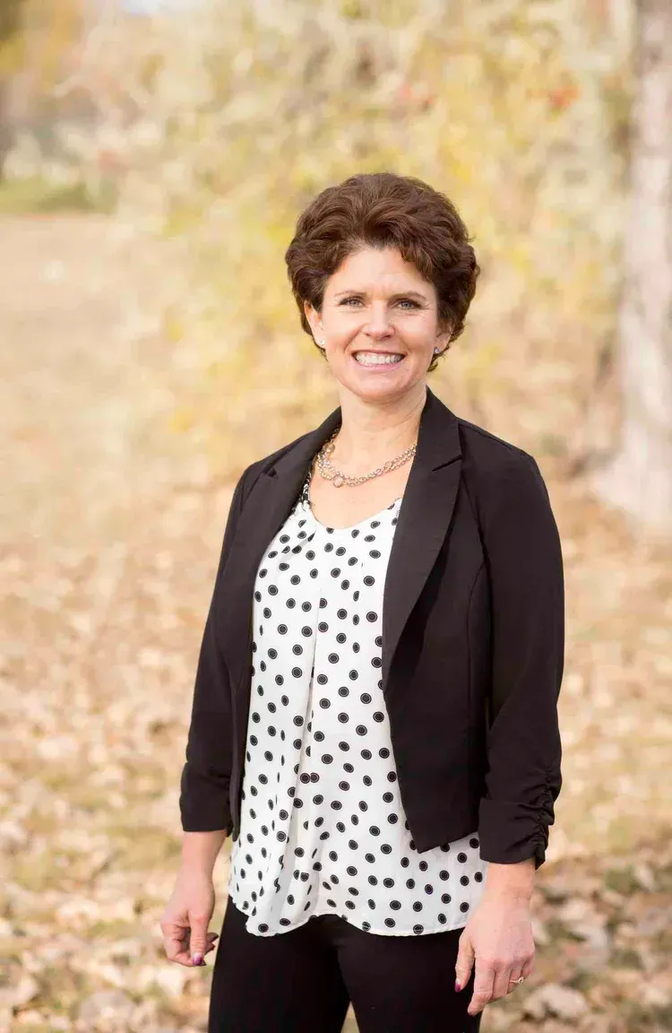 Woman with curly brown hair smiles, wearing black blazer and polka-dot shirt, outdoors in fall setting.
