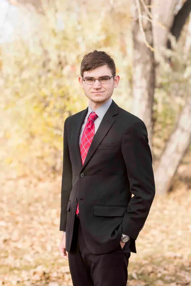 Young man in suit and tie standing outdoors, fall foliage background.