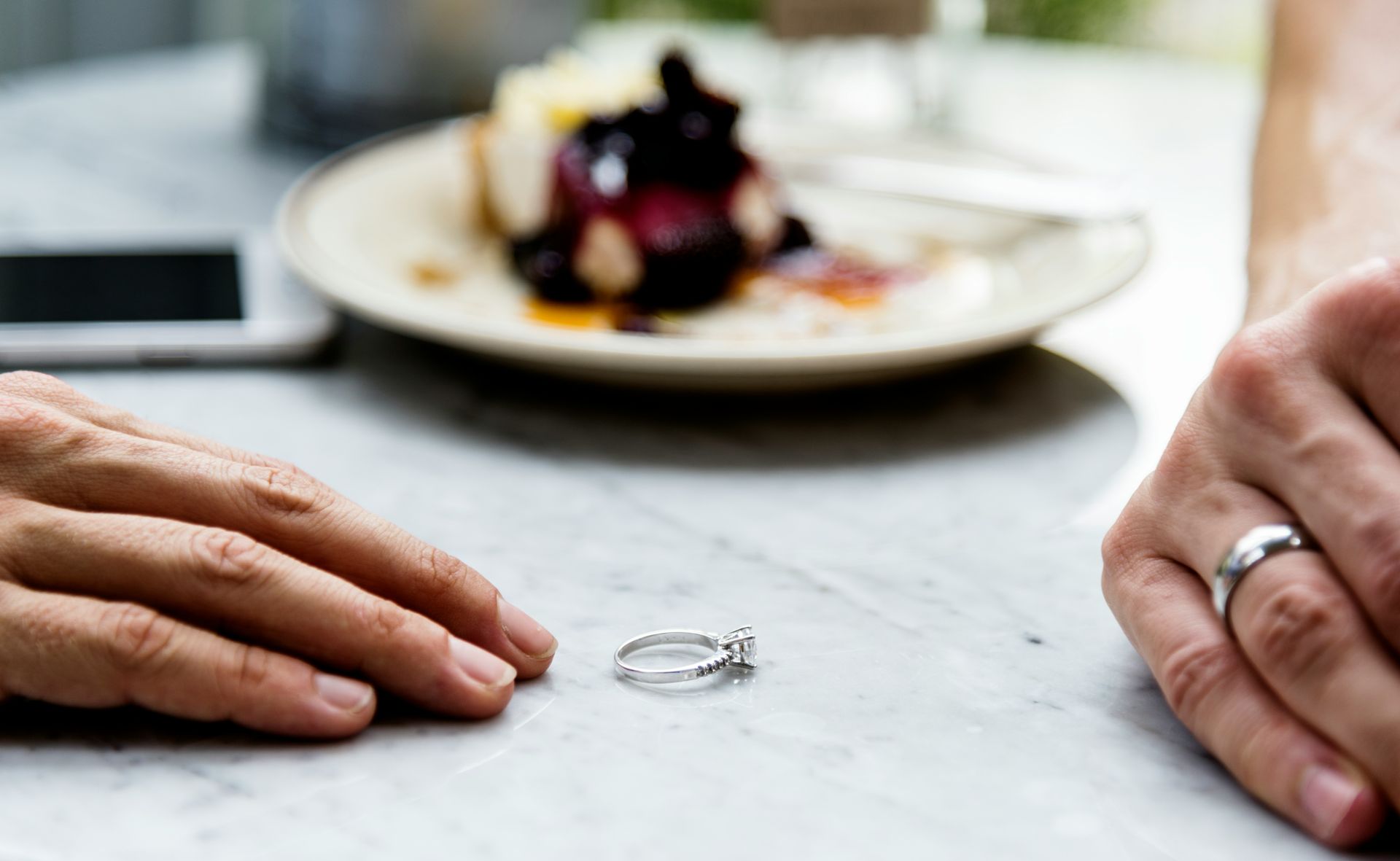 Hands near engagement ring on table; plate with dessert in background.