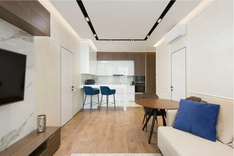 Interno di un moderno monolocale con cucina bianca, tavolo da pranzo, divano beige con cuscino blu e mobile TV.