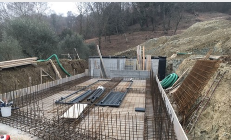 Cantiere edile che mostra le fondamenta di una piscina in cemento armato con barre di armatura metallica, tubature idrauliche e pendii di terra.