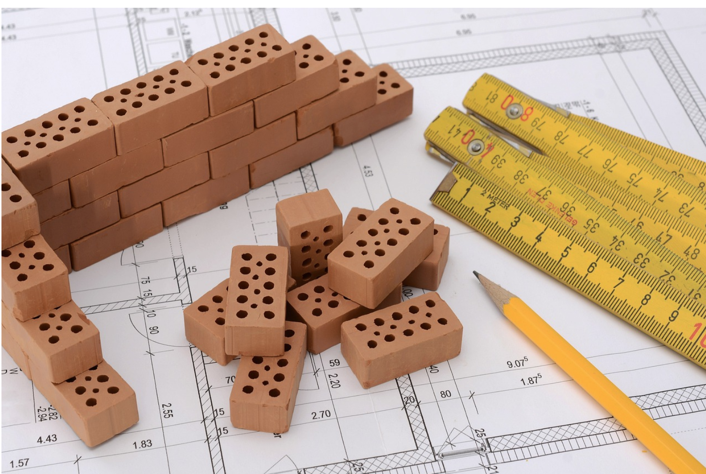 Mattoni cavi impilati, un righello pieghevole giallo e una matita poggiano su progetti architettonici.