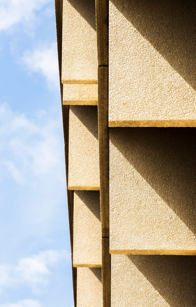 Primo piano astratto della facciata di un edificio moderno, caratterizzato da pannelli geometrici color beige con texture e ombre nette illuminate dal sole.