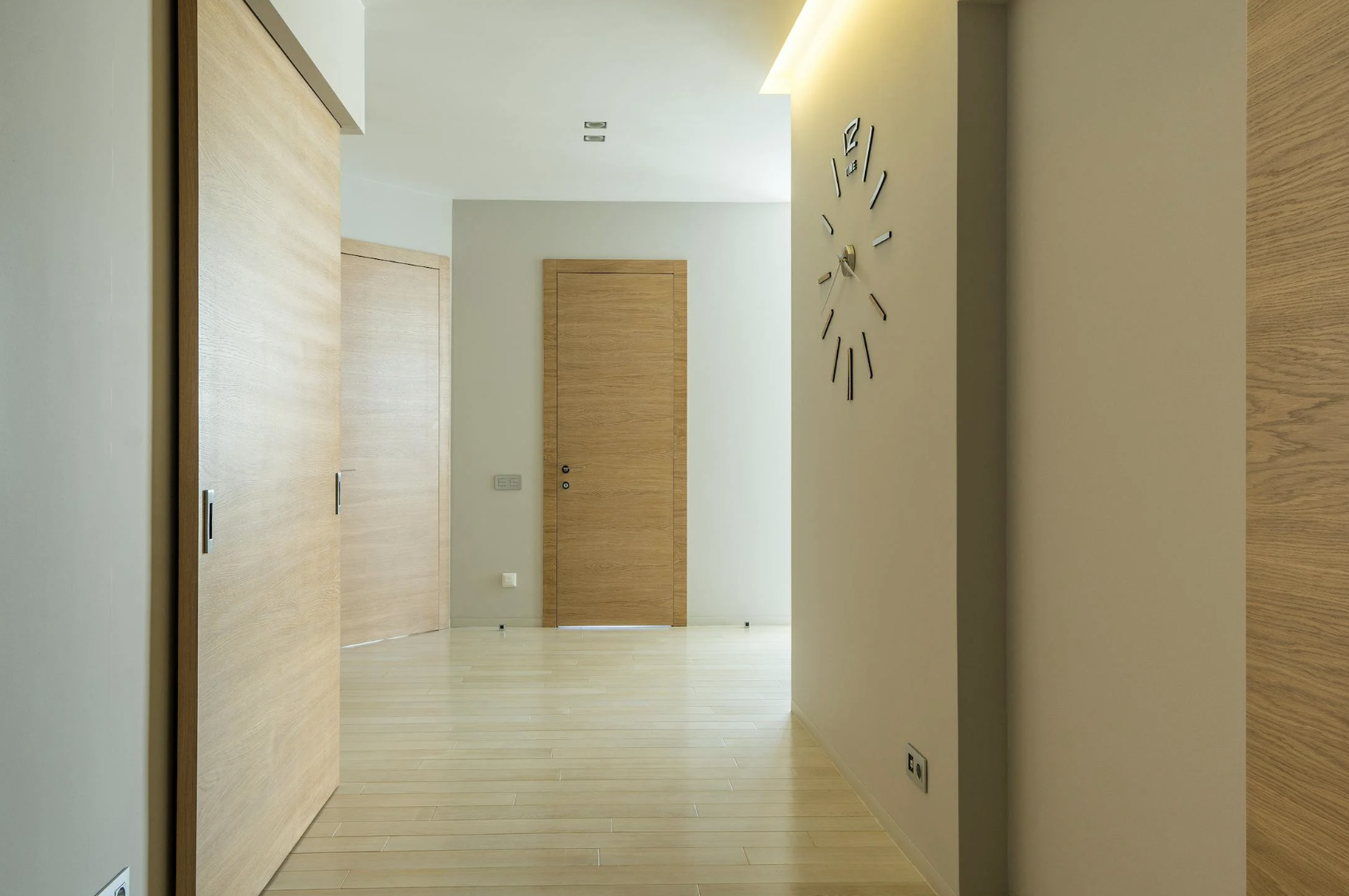 Un corridoio luminoso con pavimento in legno chiaro, pareti color beige e due porte in legno, su cui è appeso un orologio astratto in metallo.
