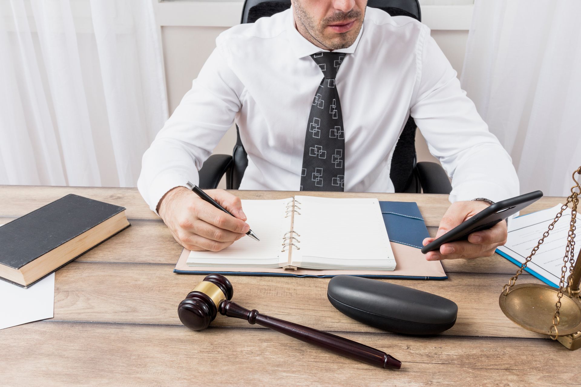 A man is sitting at a desk holding a cell phone and writing in a notebook.
