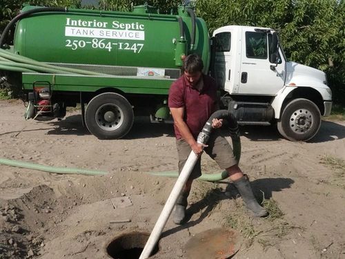 A man is pumping water into a septic tank in front of a green interior septic tank service truck