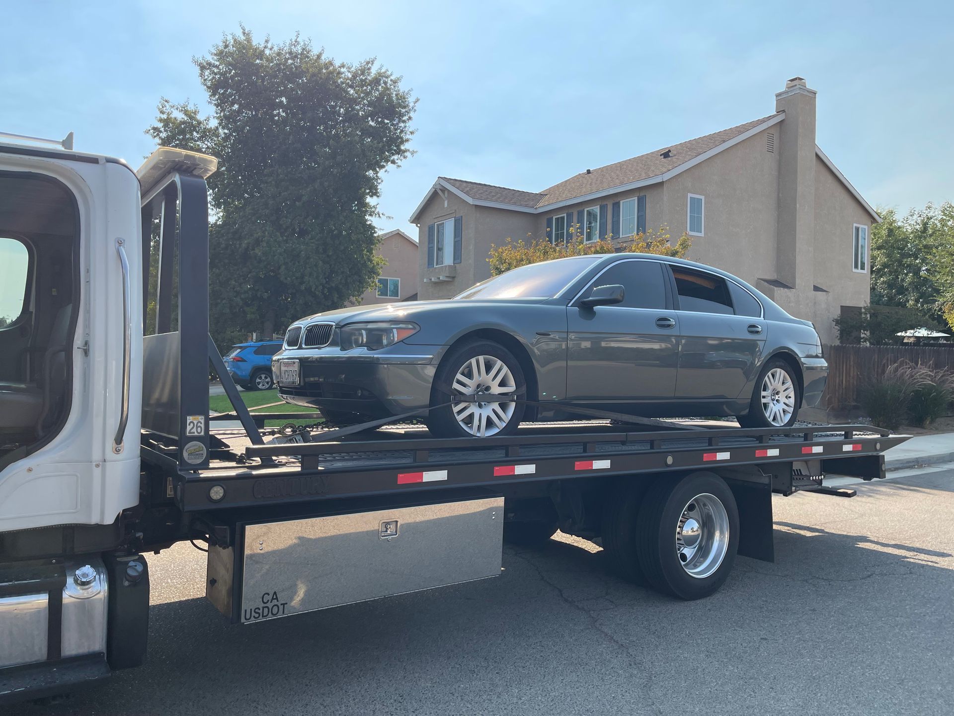 A gray BMW sedan being towed on a flatbed tow truck in front of a house.