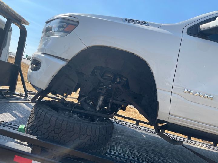White pickup truck on a tow truck, missing its front tire. Sunny outdoor setting.