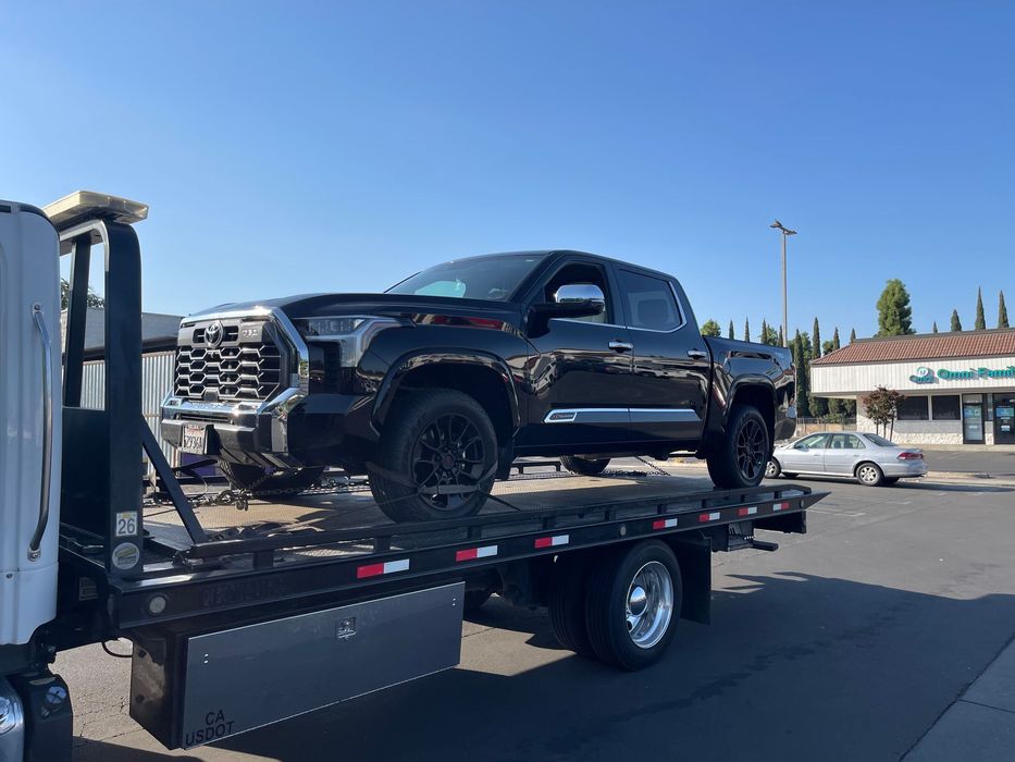 Black Toyota truck loaded on a flatbed tow truck in a sunny outdoor setting.