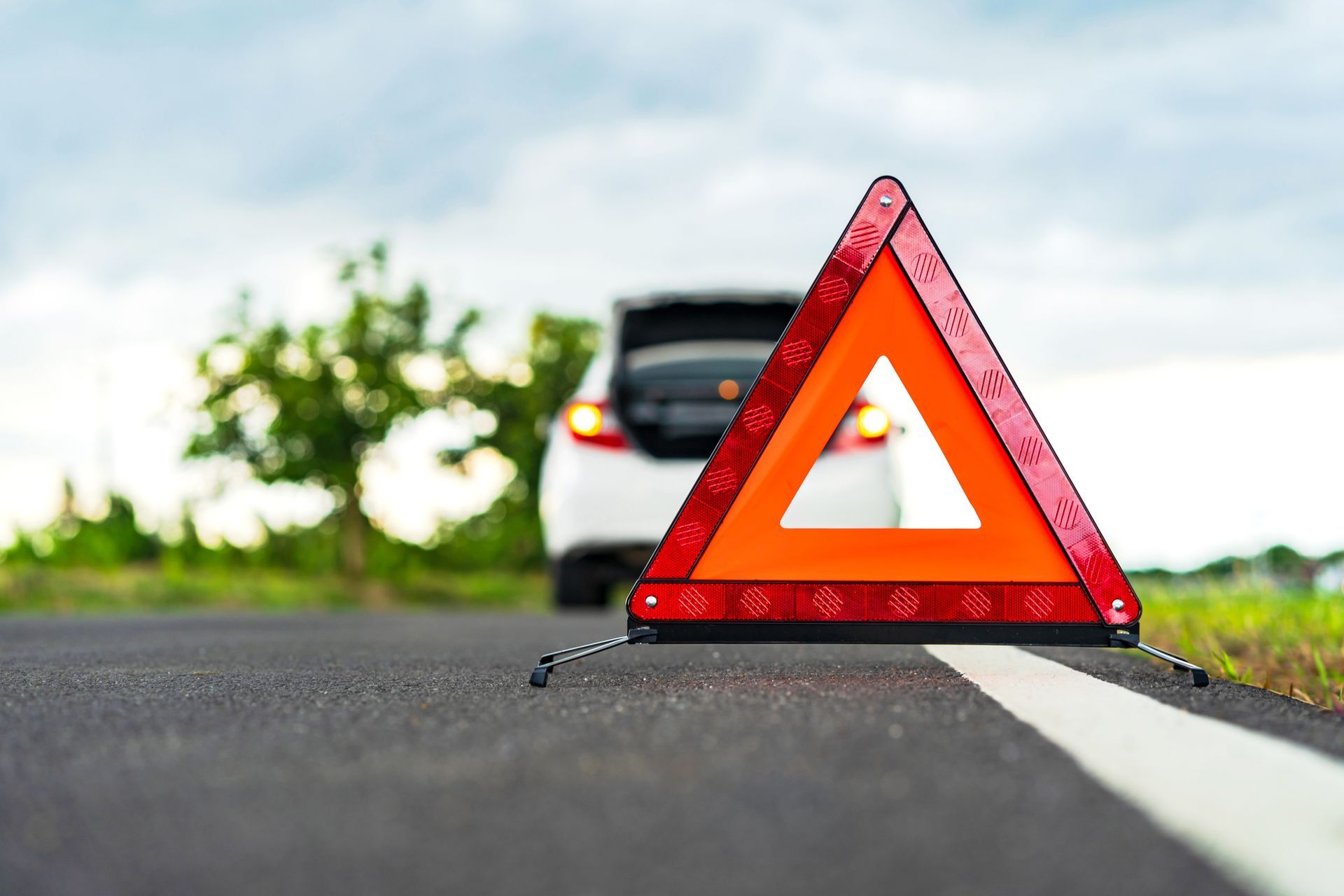 Orange warning triangle on asphalt road, car with open trunk in background.