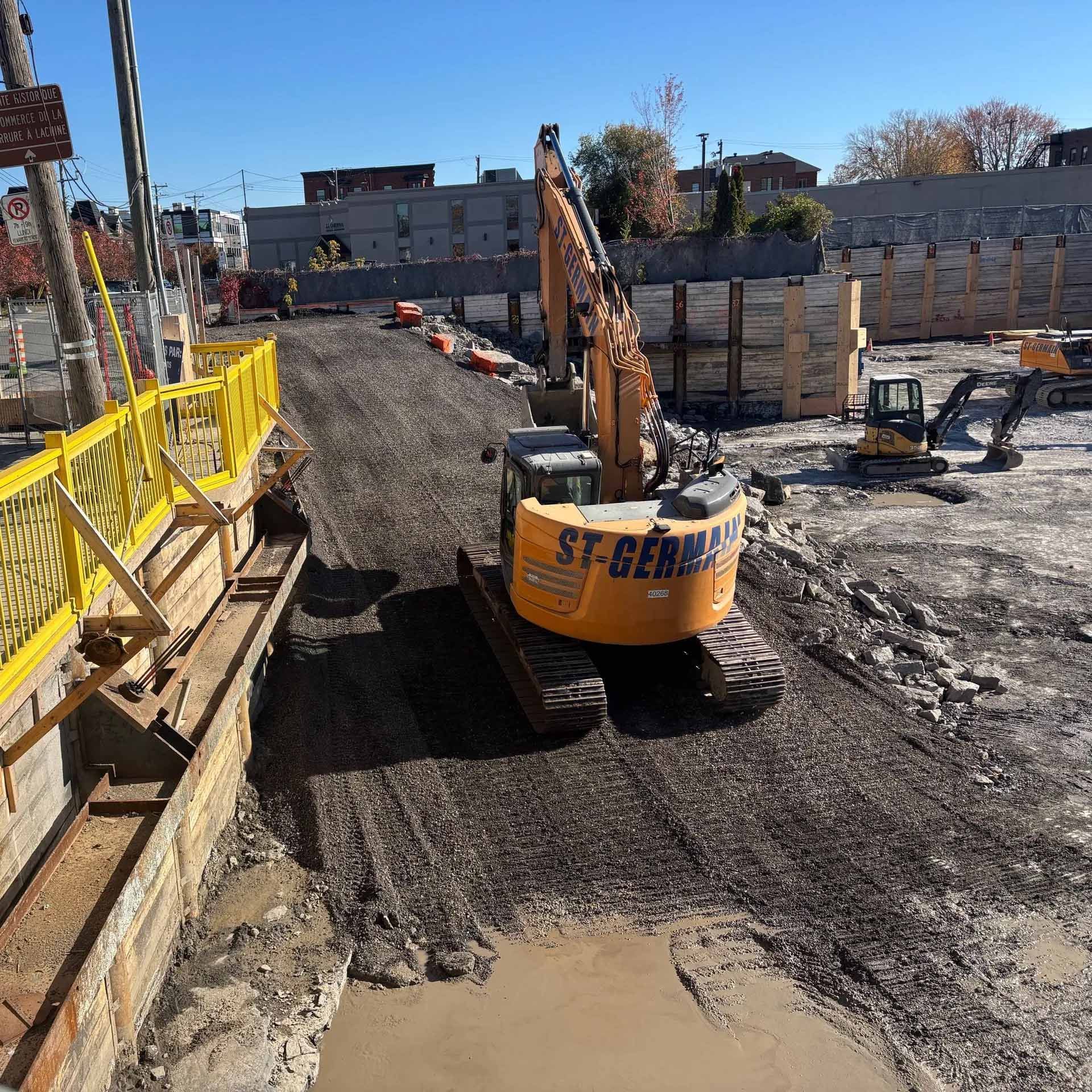 Une pelle mécanique jaune St-Germain travaille sur un chantier de construction avec des chemins de terre, des murs de soutènement et d'autres équipements.
