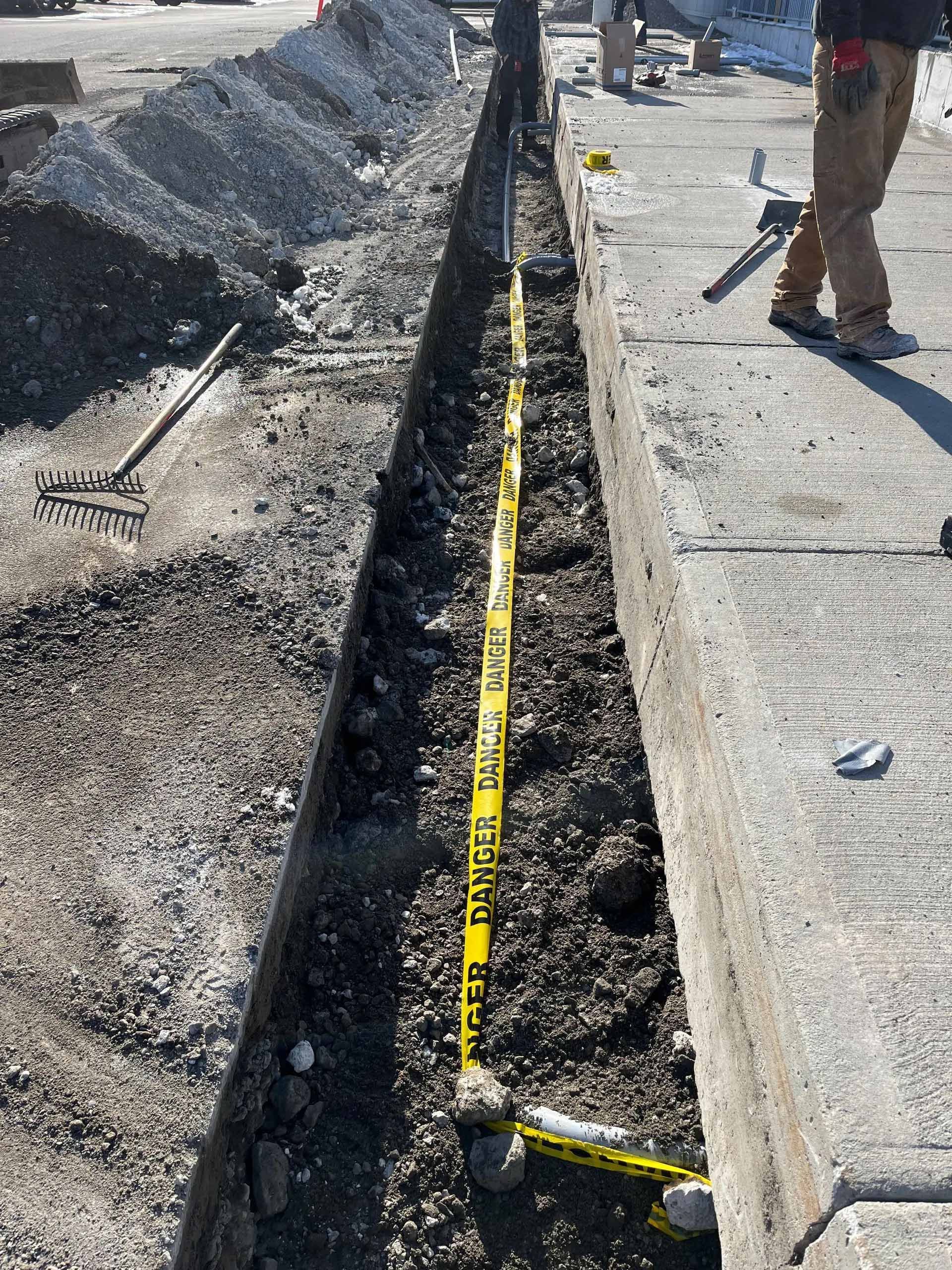 Un ouvrier se tient près d'une tranchée étroite remplie de terre, signalée par un ruban de signalisation jaune « DANGER », sur un chantier de construction.