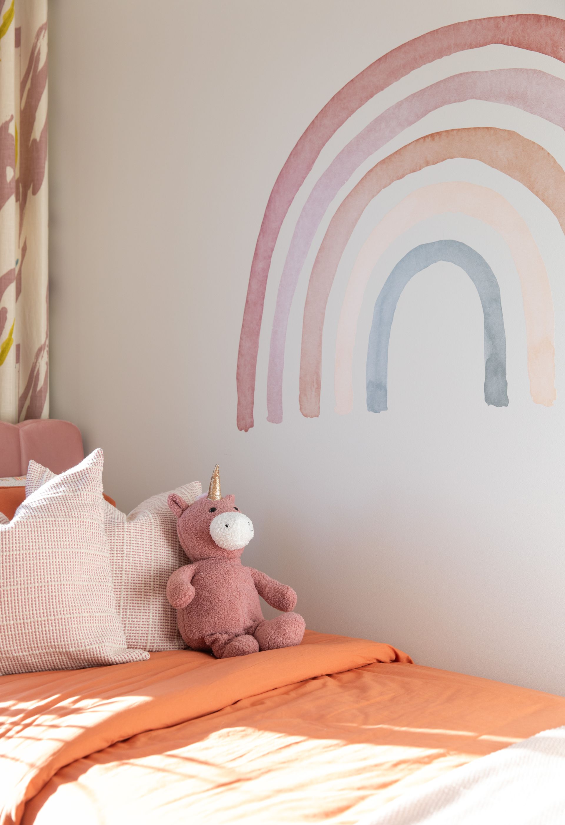 A stuffed unicorn is sitting on a bed next to a rainbow on the wall.