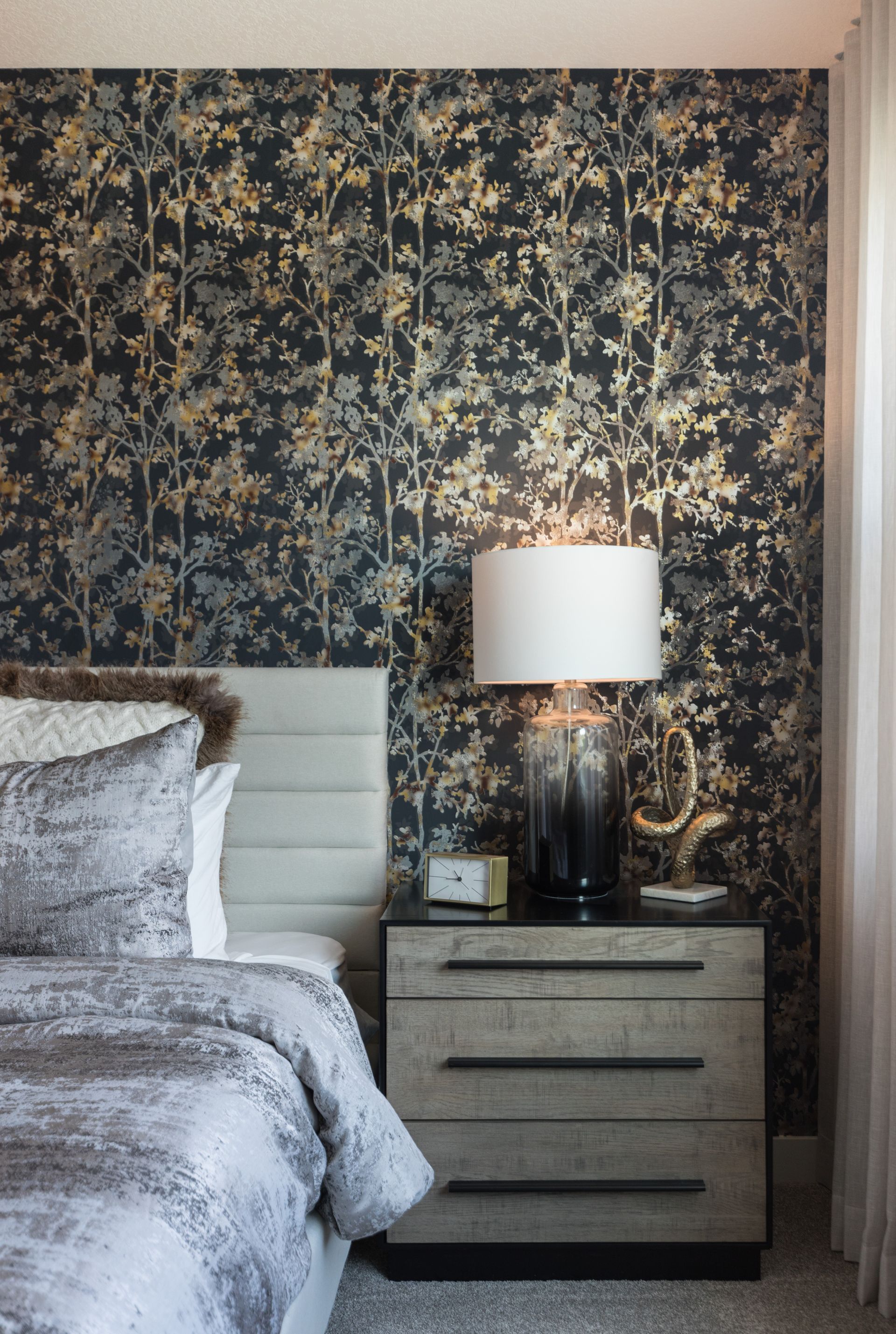 A bedroom with a bed , nightstand , lamp and wallpaper.