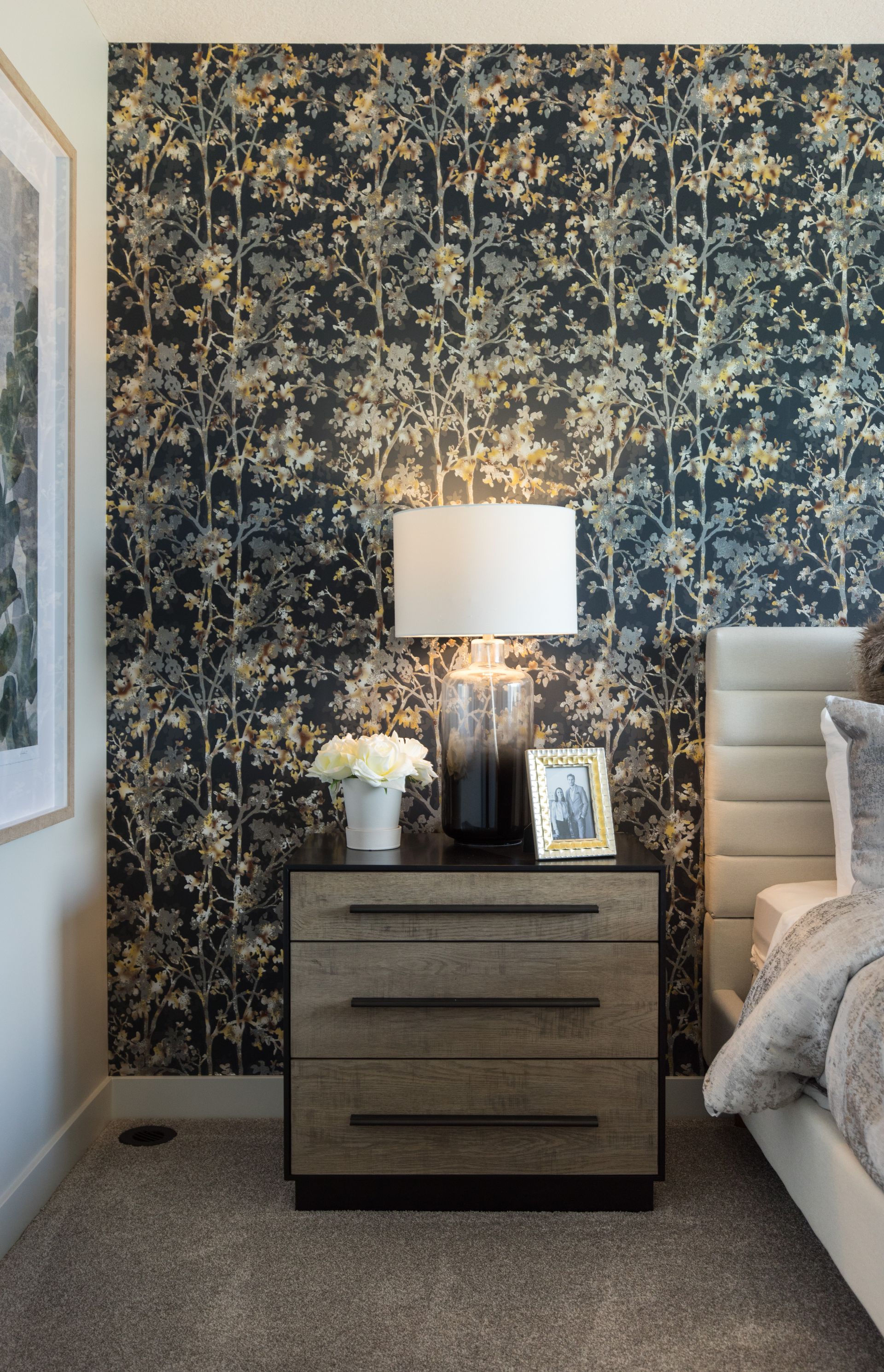A bedroom with a bed , nightstand , lamp and floral wallpaper.
