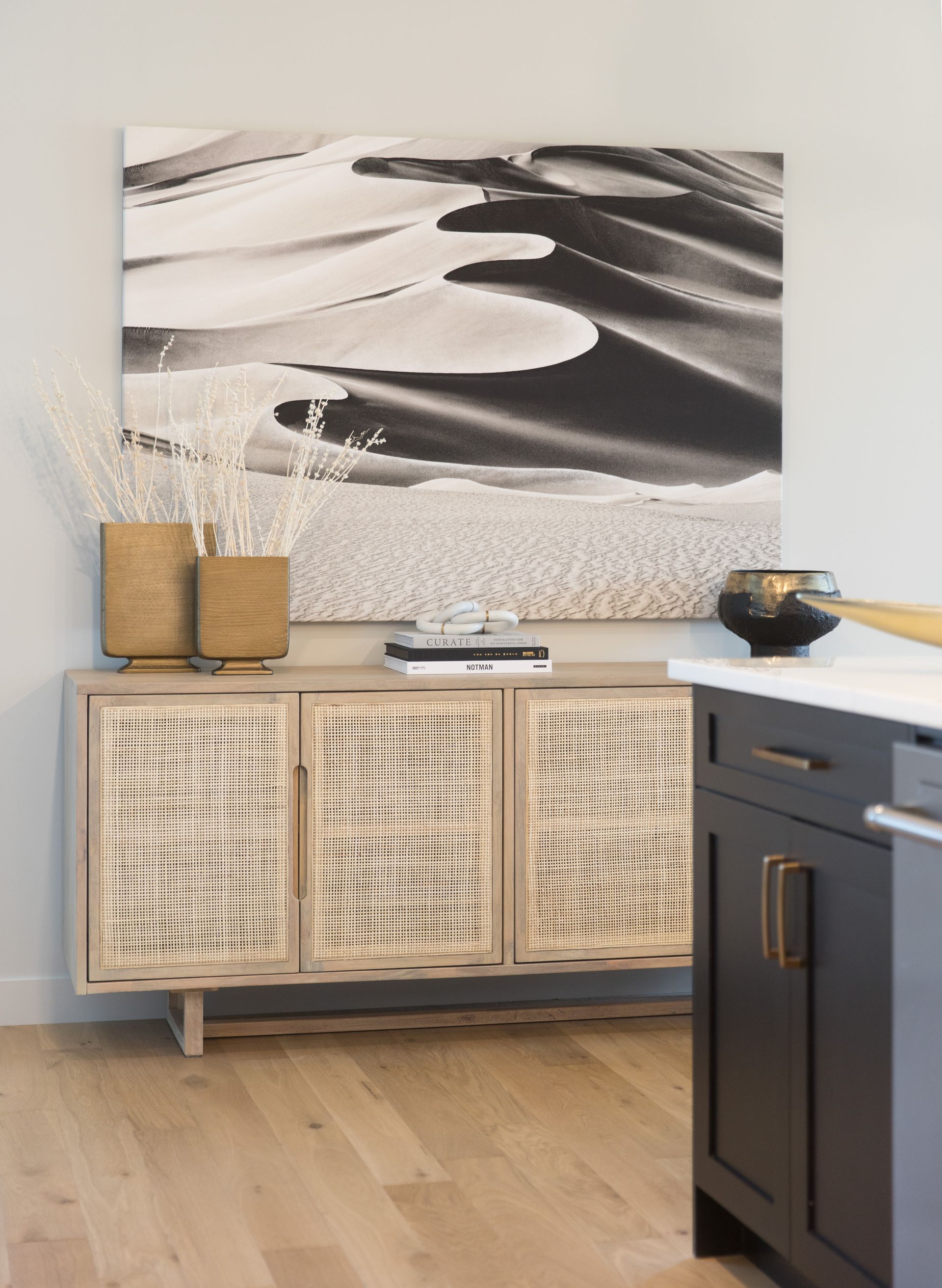 A black and white painting hangs above a wicker cabinet in a kitchen.