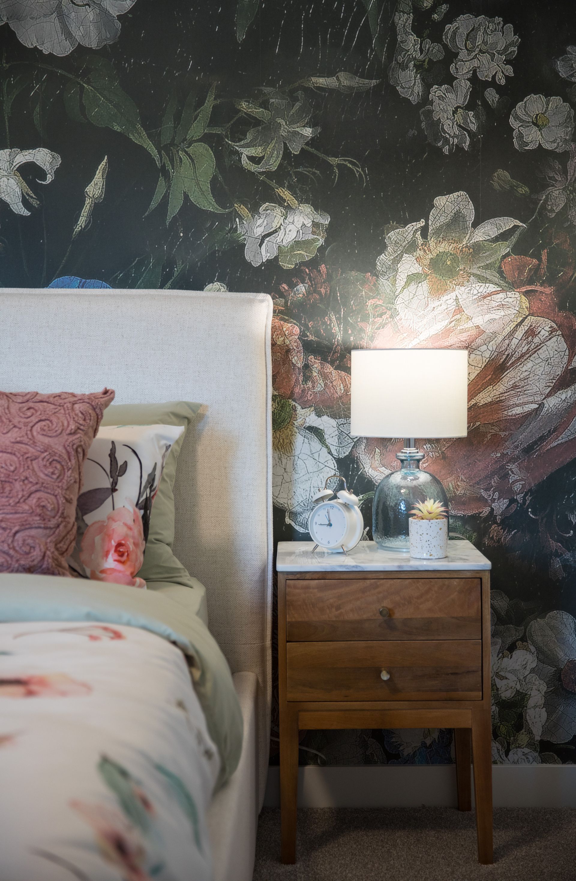 A bedroom with a bed , nightstand , lamp and floral wallpaper.