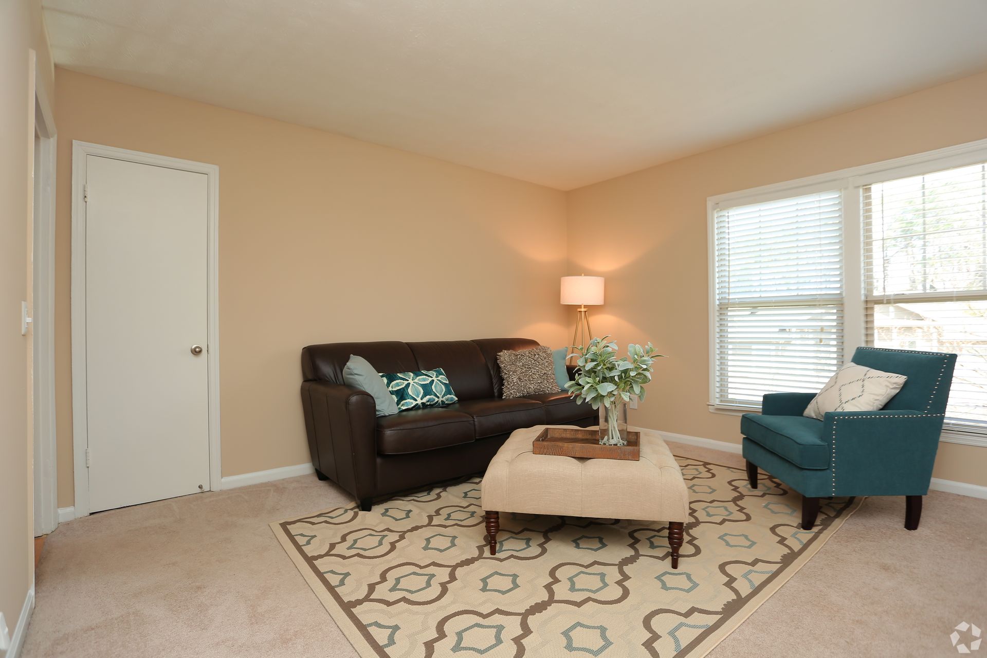 Bright living room with brown sofa, teal chair, patterned rug, coffee table, and large windows.