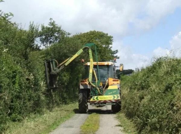 Tractor with a hedge trimmer clearing a narrow, overgrown country lane.