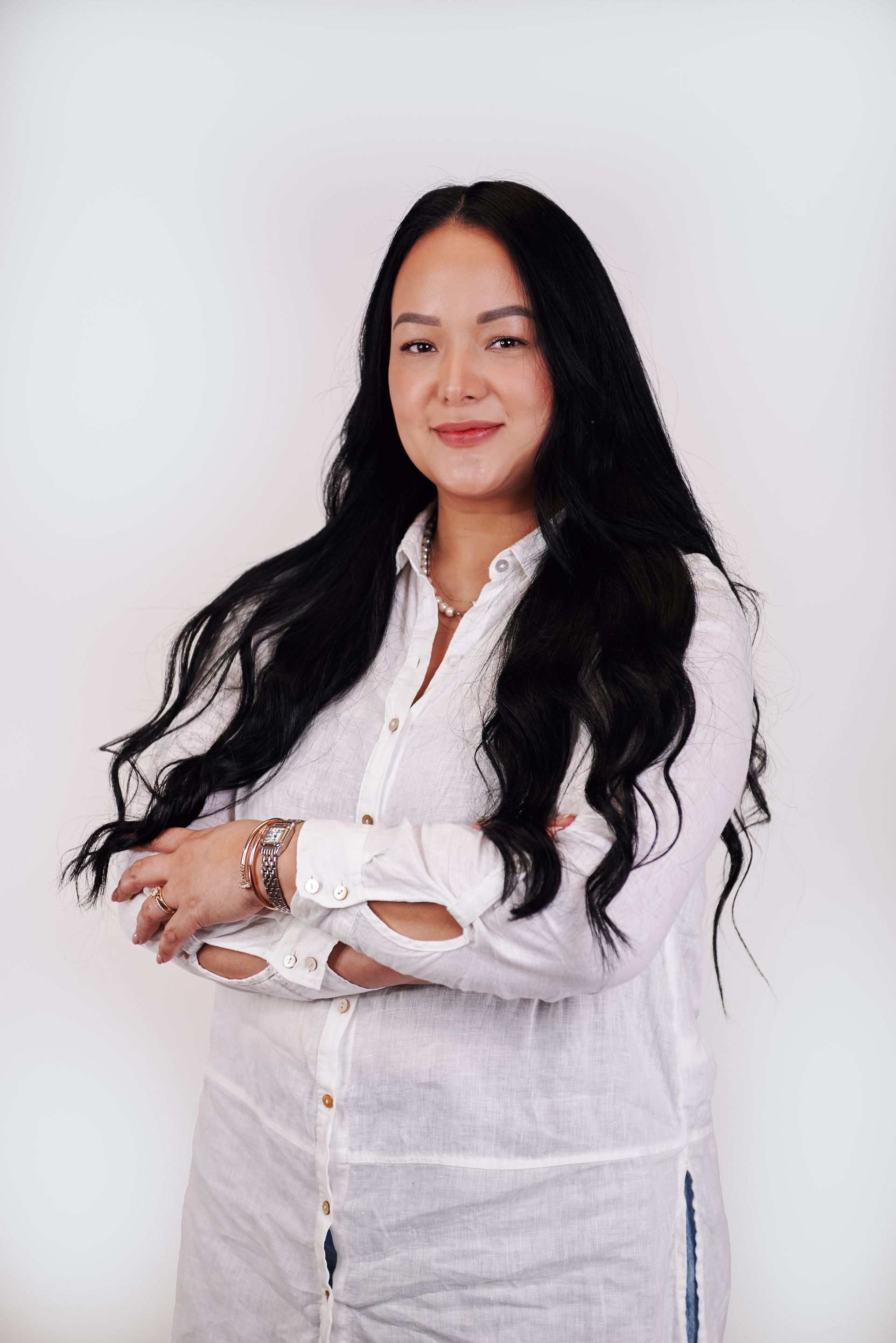 A woman with long black hair is wearing a white shirt and standing with her arms crossed.