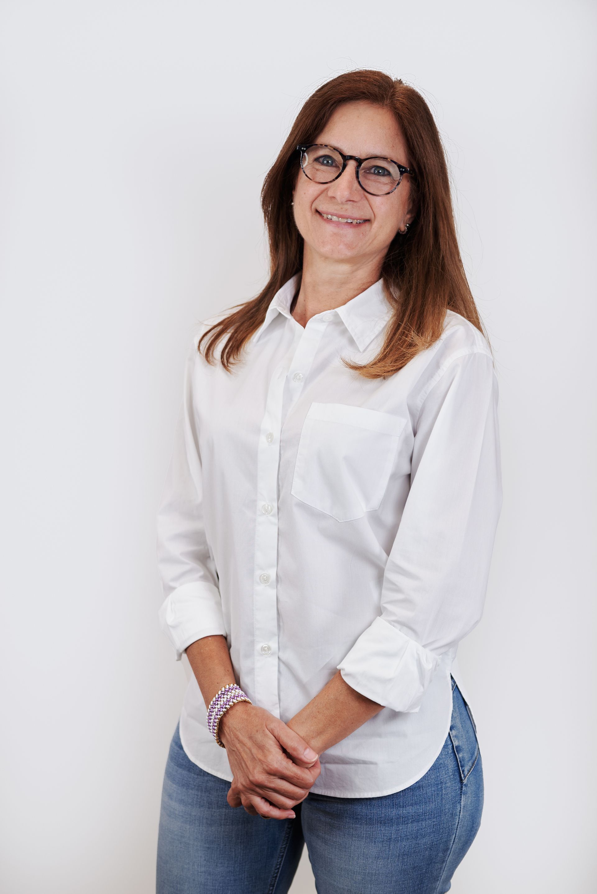 A woman wearing glasses and a white shirt is standing in front of a white wall.