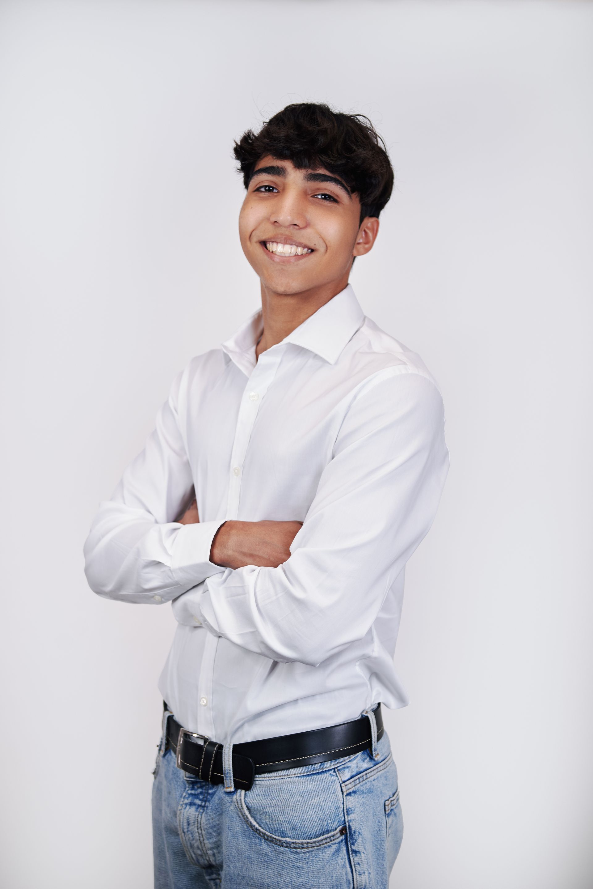 A young man in a white shirt and blue jeans is standing with his arms crossed and smiling.