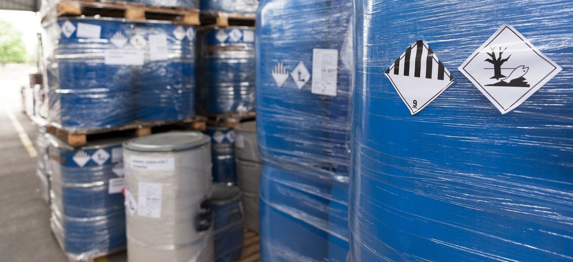 Des fûts de produits chimiques bleus portant des symboles de danger sont empilés dans un entrepôt.