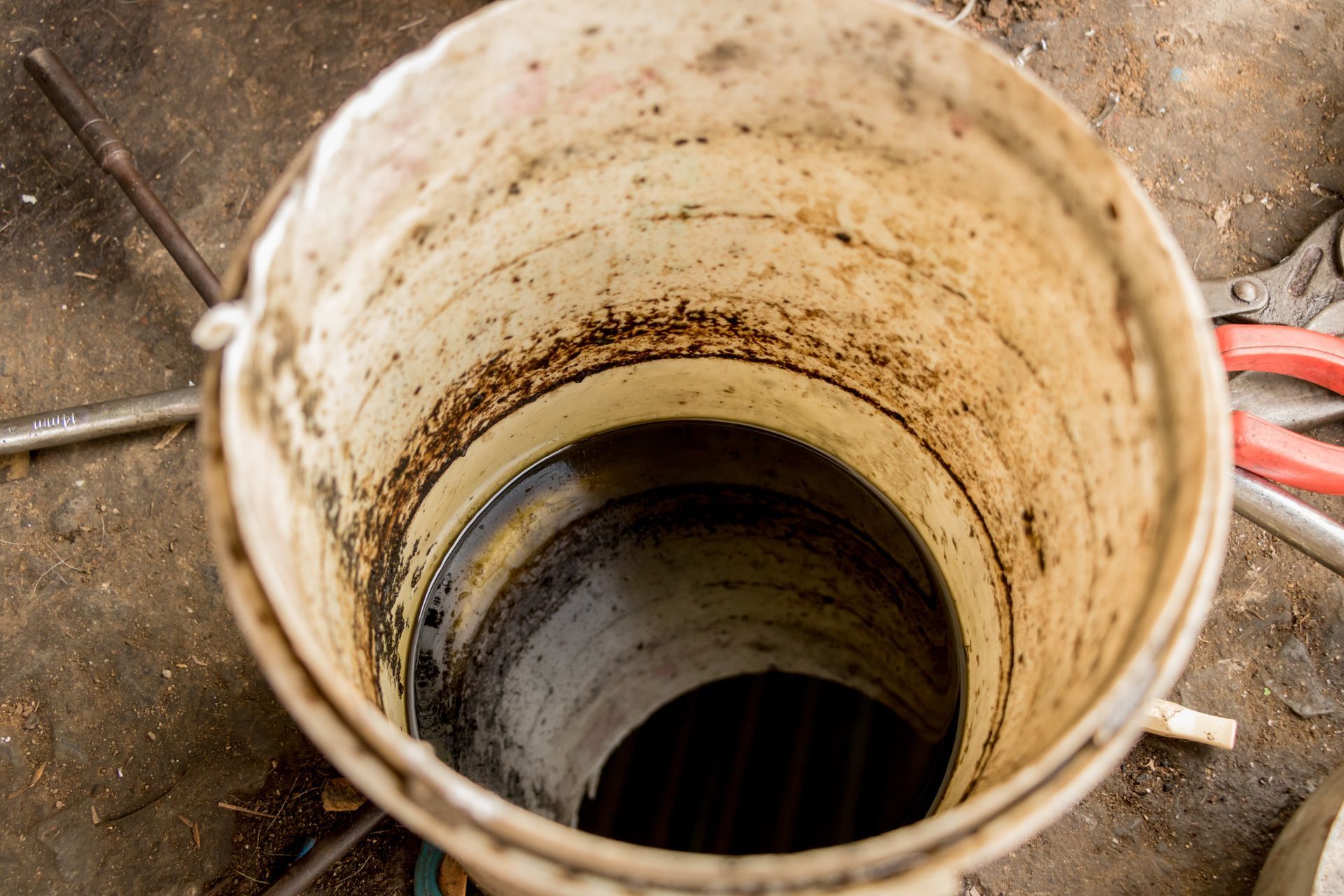 Seau blanc et sale rempli de liquide foncé, vu d'en haut, entouré d'outils sur une surface en béton.