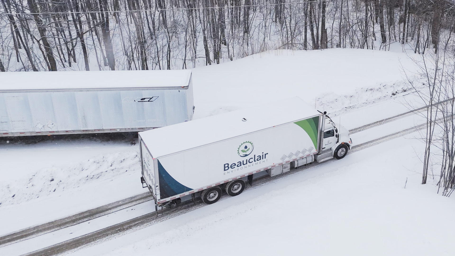 Semi-camion blanc avec remorque sur une route enneigée, dépassant un autre camion, arbres en arrière-plan.