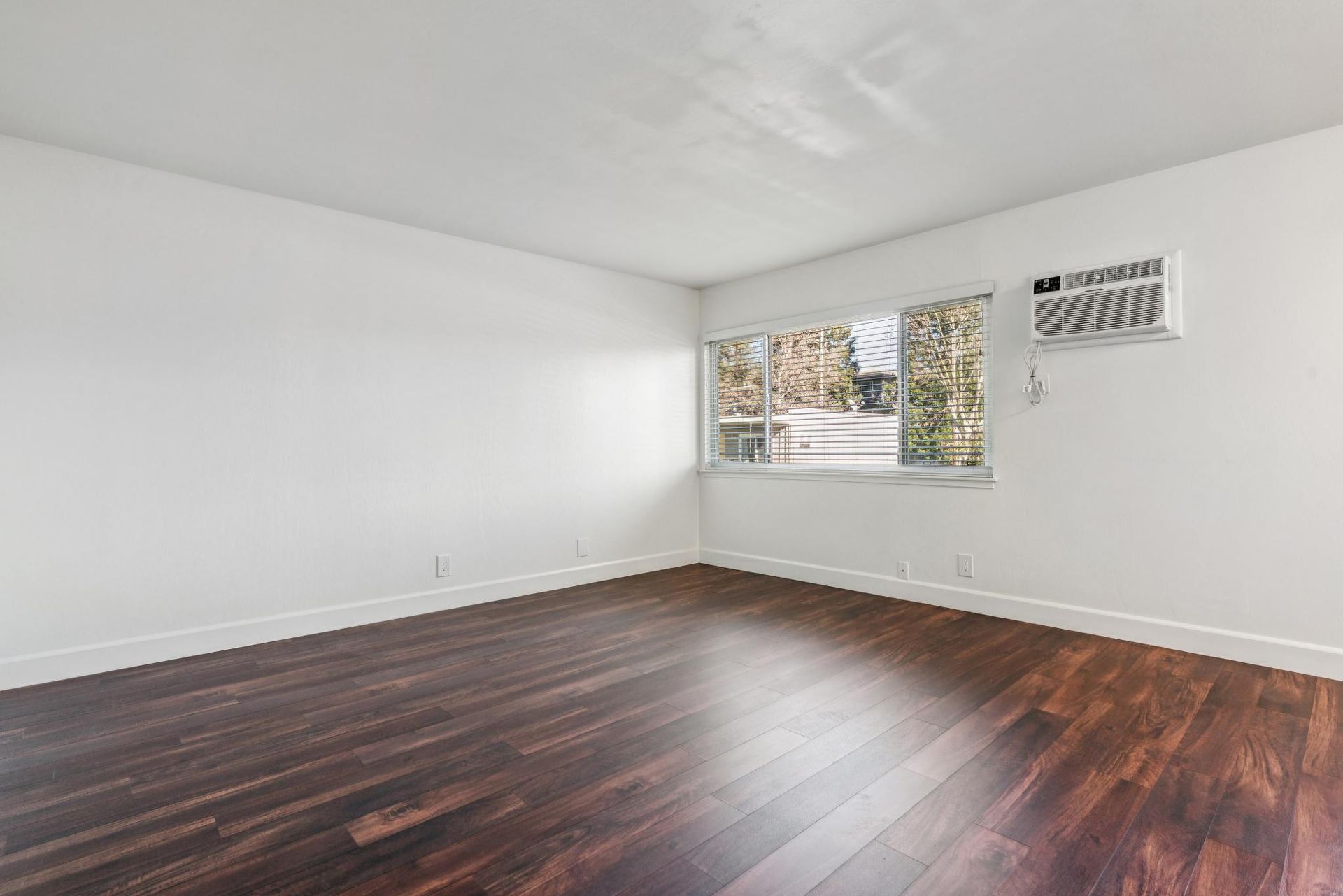 Empty room with dark wood floor, white walls, and a window with an air conditioner.
