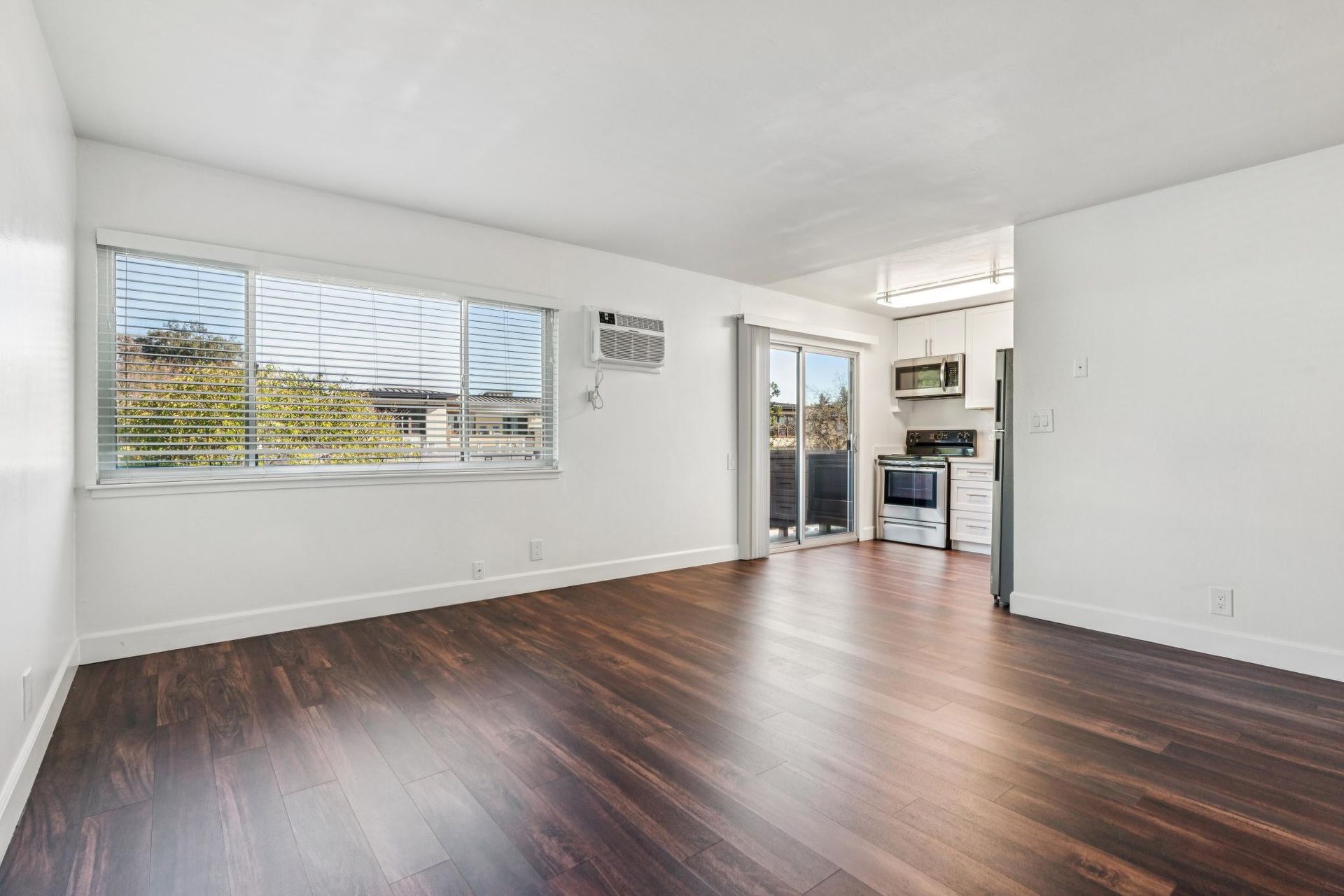 Empty living room with dark wood floors, large windows, and open kitchen.