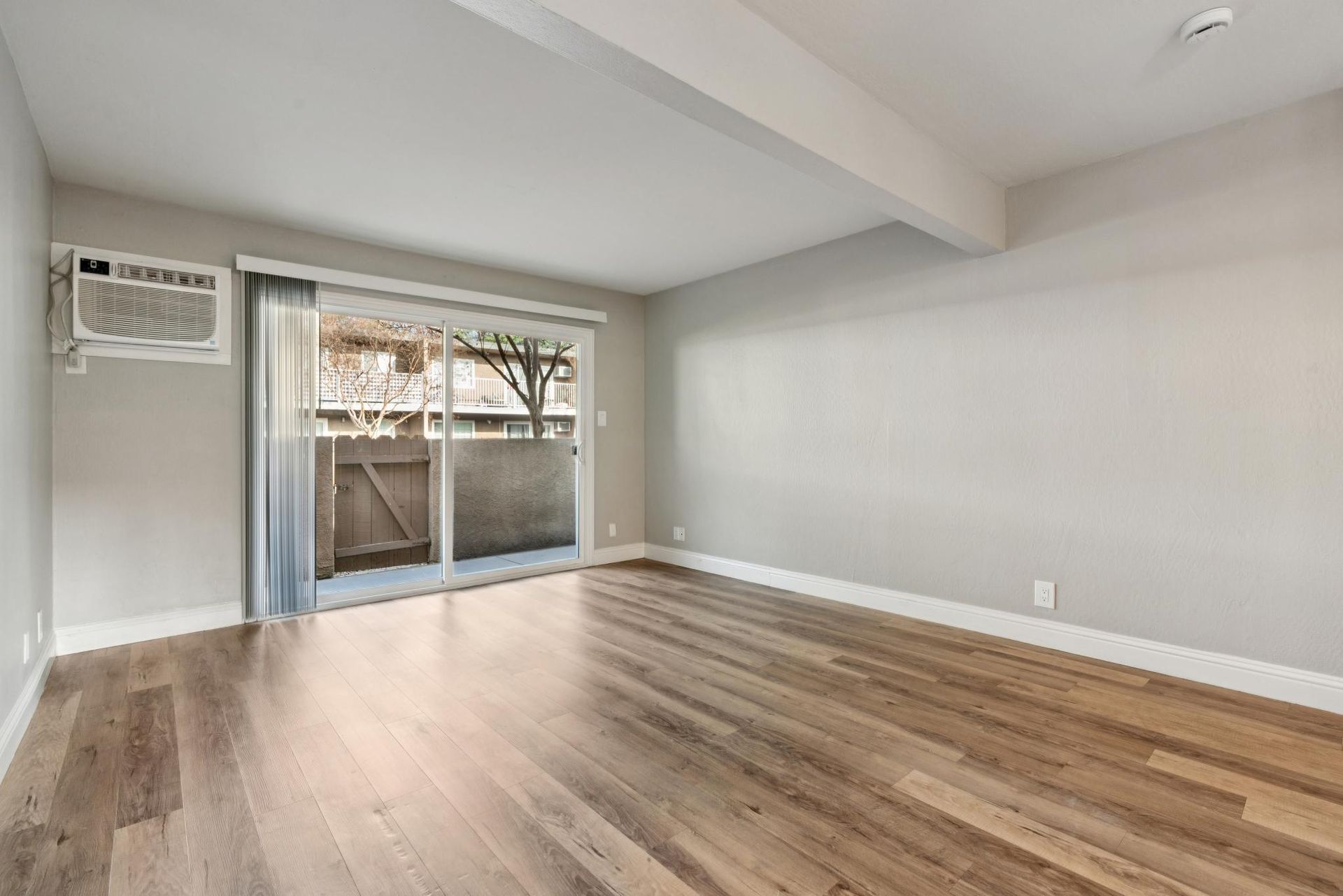 Empty living room with wood flooring, sliding glass door to a balcony, and air conditioning unit.