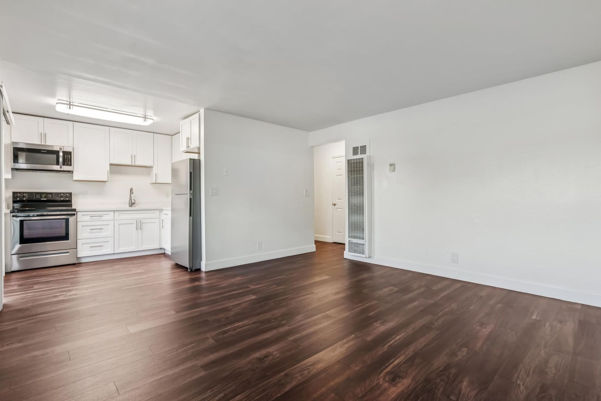 Empty apartment interior with white walls, dark wood floors, and stainless steel appliances in the kitchen.